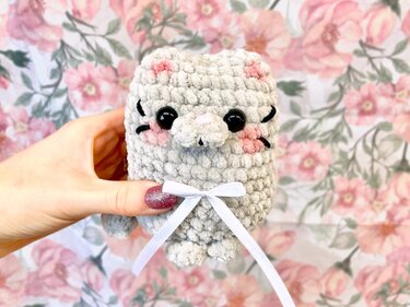 Hand hält graue gehäkelte Amigurumi-Katze mit schwarzen Augen, rosa Bäckchen und weißer Schleife vor floralem Hintergrund