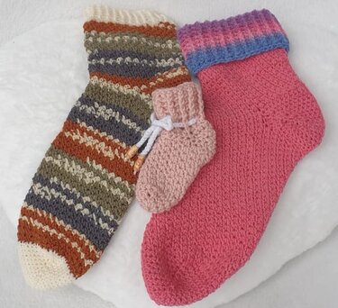 Drei gehäkelte Socken: eine lange gestreifte Socke, eine pinke Socke mit mehrfarbigem Bund und eine kleine rosa Babysocke mit Kordel.