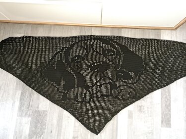 Schwarzes, dreieckiges, gehäkeltes Tuch mit Hundegesicht-Motiv in Filethäkeltechnik auf hellem Boden ausgebreitet.