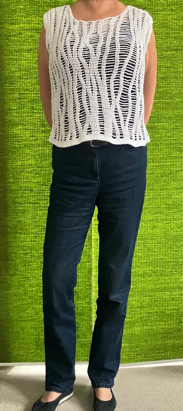 Person trägt weißes, ärmelloses Häkeloberteil mit durchbrochenem Wellen-Ajourmuster über dunklem Top und dunklen Jeans vor grünem Hintergrund.