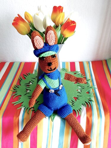Brauner Amigurumi-Hase mit blauer Latzhose, Halstuch und Mütze vor Tulpenvase auf bunt gestreifter Tischdecke.