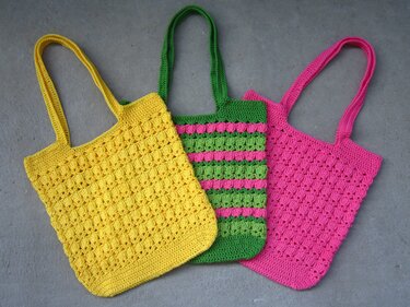 Drei gehäkelte Shopper in Gelb, Grün und Pink auf grauem Hintergrund
