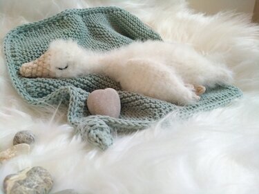 Gehäkelter weißer Schwan mit beigem Schnabel und geschlossenen Augen auf mintgrüner Strickdecke und weißem Plüsch.