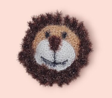 Gehäkelter Löwenkopf mit dunkelbrauner, buschiger Mähne und hellem Gesicht auf rosa Hintergrund