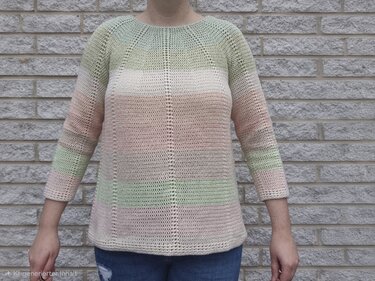 Gehäkelter Damenpullover in Pastellgrün, Creme und Rosé mit Dreiviertelärmeln vor einer Ziegelwand