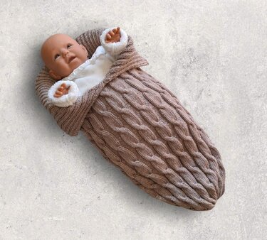 Babypuppe in gestricktem taupefarbenem Schlafsack mit Zopfmuster, umlegbarem Kragen und flauschigen Bündchen.