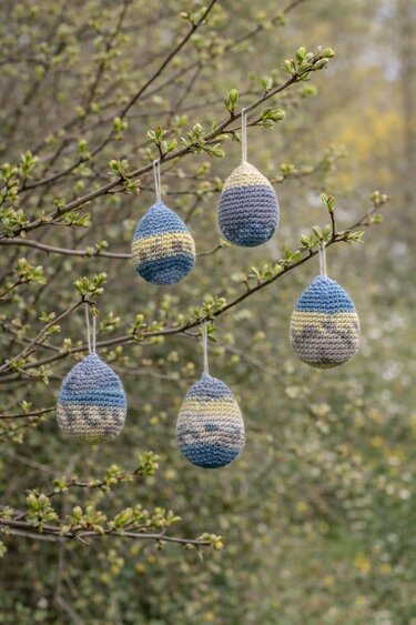Fünf gehäkelte Ostereier in Blau und Creme an einem Zweig mit Knospen