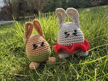 Zwei gehäkelte Osterhasen in Eiform im grünen Gras, einer braun-grün und einer beige mit roter Rüsche.