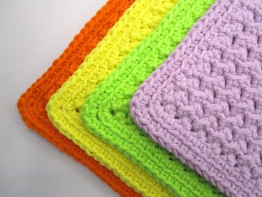 Vier übereinandergelegte gehäkelte Topflappen in Orange, Gelb, Neongrün und Lila, Nahaufnahme der Maschenstruktur