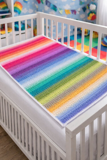 Bunte gehäkelte Regenbogen-Babydecke ausgebreitet in einem weißen Gitterbett