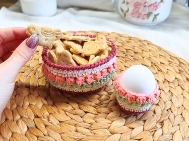 Gehäkeltes Körbchen mit Tulpenmuster, gefüllt mit Keksen in Tierform, daneben ein passender gehäkelter Eierbecher mit weißem Ei auf einem geflochtenen Platzset.