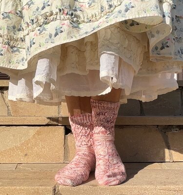 Beine mit rosa gestrickten Socken mit Zopfmuster und Rippenbund unter mehrlagigem Rock, stehend auf Steintreppe