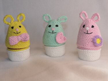Drei gehäkelte Osterhasen-Eierwärmer: gelb mit rosa Schleife, mintgrün mit rosa Herz, rosa mit lila-gelber Blume auf weißen Sockeln.