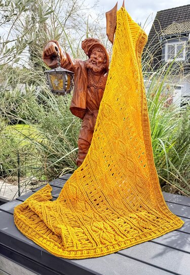 Golden yellow crochet triangle shawl with cable and lace patterns draped over a wooden garden statue outdoors.