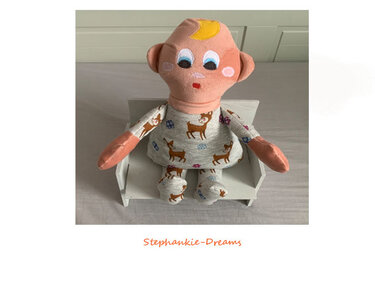 Handmade plush baby doll with a peach head, yellow crescent, embroidered face, and gray deer-patterned outfit sitting on a small white bench.