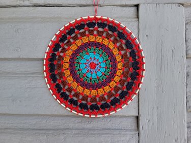 Rundes gehäkeltes Mandala mit bunten Ringen, rotem Rand und türkisem Zentrum, an einer grauen Holzwand hängend.