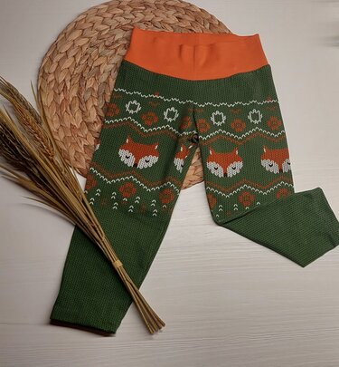 Schmale grüne Kinderleggings mit hohem orangefarbenem Bündchen und Fuchsmotiven, auf hellem Holz mit geflochtener Matte und Getreidebündel.