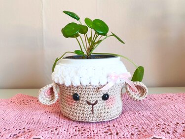 Gehäkelter Schaf‑Übertopf in Beige und Weiß mit rosa Schleife und grüner Pflanze auf rosa Häkeldecke.