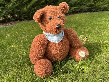 Brauner, flauschig gestrickter Teddybär mit hellblauem Dreieckstuch sitzt im Gras neben einem Gänseblümchen.