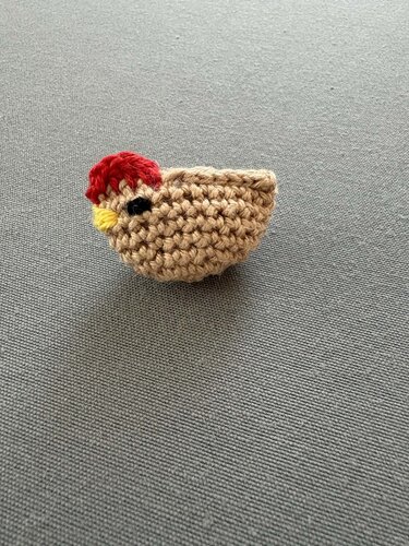Gehäkeltes Küken-Amigurumi in Beige mit rotem Kamm und schwarzem Auge auf grauem Untergrund.