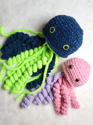 Zwei gehäkelte Quallen: dunkelblaue mit neongrünen Tentakeln und pinke mit lavendelfarbenen Tentakeln auf hellem, flauschigem Untergrund.