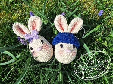 Zwei gehäkelte Hasen in Eierform im Gras, einer mit lila Schleife, einer mit blauer Mütze.
