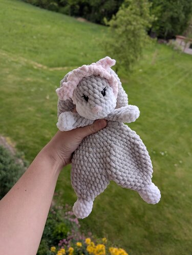 Hand hält gehäkeltes Häschen-Schmusetuch in Grau, Weiß und Rosa vor grüner Wiese