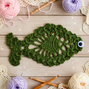 Green crocheted fish-shaped coaster with craft eye on wooden tabletop