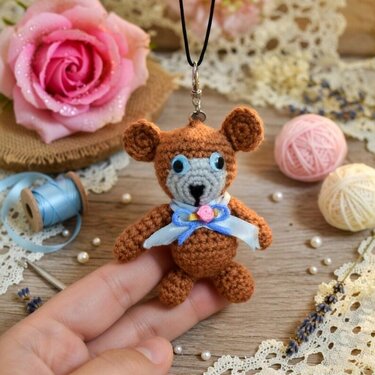Small brown crochet teddy bear keychain held in hand, surrounded by crafting supplies