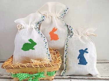Drei weiße Stoffbeutel mit Kordelzug, bestickt mit grünen, orangefarbenen und blauen Hasen im Kreuzstich; ein Beutel steht in einem geflochtenen Korb mit Stroh.