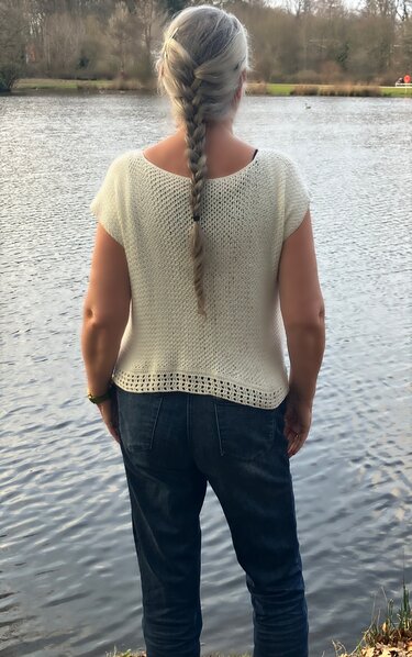 Rückenansicht: Person trägt cremefarbenes, kurzärmeliges Häkelshirt mit Strukturmaschen und offener Saumkante