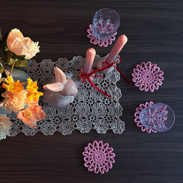 Vogelperspektive eines dunklen Holztisches mit grauem gehäkeltem Tischläufer, rosa Kerzen, rosa gehäkelten Untersetzern, Gläsern, Hasenfigur und Blumen.