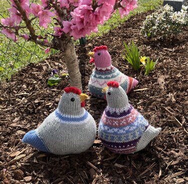 Drei gestrickte Hühner-Amigurumi mit bunten Streifen unter rosa Blüten