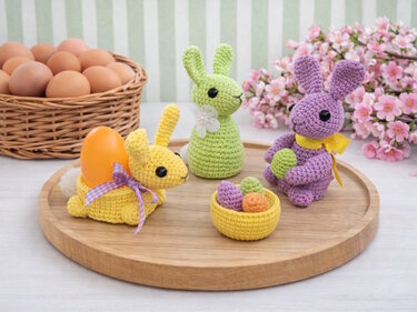 Drei gehäkelte Amigurumi-Hasen in Gelb, Grün und Lila mit Eiern auf einem runden Holztablett.