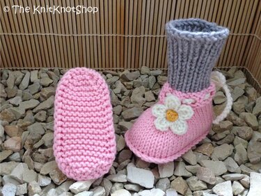 Zwei rosa gestrickte Babyschuhe, einer liegend mit sichtbarer krauser Sohle, einer mit grauem Rippenschaft und weißer Blütenapplikation