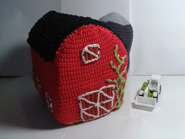 Nahaufnahme eines roten, gehäkelten Bauernhauses mit schwarzem Dach, grauem Silo und aufgestickten Ranken