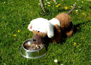 Gehäkelter brauner Amigurumi-Hund mit weißen Ohren neben Metall-Futternapf auf Gras