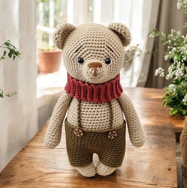 Beiger gehäkelter Teddybär mit geripptem dunkelrotem Schal und dunkler Latzhose auf Holztisch.