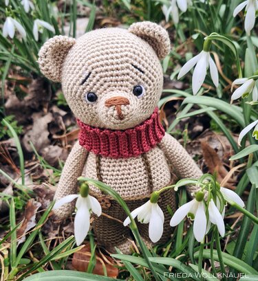 Beiger gehäkelter Amigurumi-Teddybär mit rotem Schal und brauner Hose zwischen Schneeglöckchen