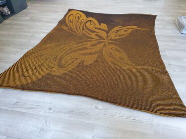 Gestrickte Decke in Braun und Senfgelb mit schmalen Streifen und großem floralem Motiv, flach auf hellem Holzboden ausgebreitet