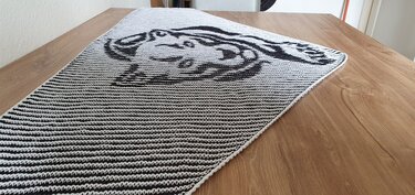 Black and white knitted shawl with a stylized creature motif on one end, laid flat on a wooden table.