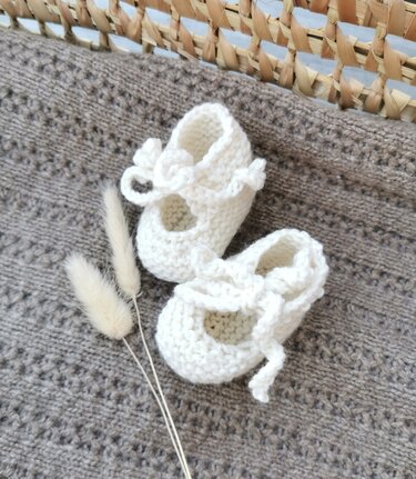 Weiße gestrickte Ballerina-Babyschuhe mit Bindeband auf brauner Strickdecke und getrockneten Gräsern