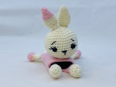 Cream crocheted amigurumi bunny with pink accents wearing a pink dress, sitting on a white surface.