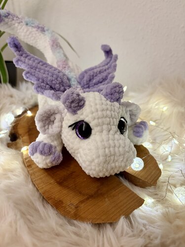 Gehäkelter weißer Drache mit fliederfarbenen Flügeln und großen dunklen Augen auf Holz- und Fellunterlage mit Lichterketten