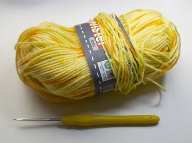 Skein of yellow and orange variegated baby yarn next to a yellow-handled crochet hook on white.