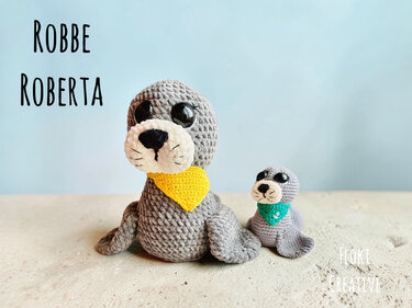 Zwei gehäkelte Robben-Amigurumi in Grau, eine größere mit gelbem Halstuch und eine kleine mit grünem Halstuch, vor hellem Hintergrund