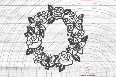 Schwarze Silhouette eines runden Blumenkranzes mit Rosen, Blüten und Schmetterlingen auf hellem Holzgrund.