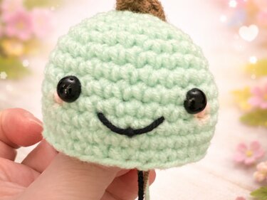 Hand hält hellgrünes gehäkeltes Amigurumi mit lächelndem Gesicht vor floral unscharfem Hintergrund