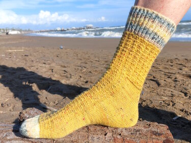 Gelbe gestrickte Socke mit mehrfarbigem Bündchen auf Treibholz am Strand, Meer im Hintergrund.