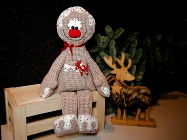 Gehäkelte Amigurumi‑Puppe mit roter Nase und weißen gepunkteten Applikationen auf einer Holzkiste neben einem Holzrentier